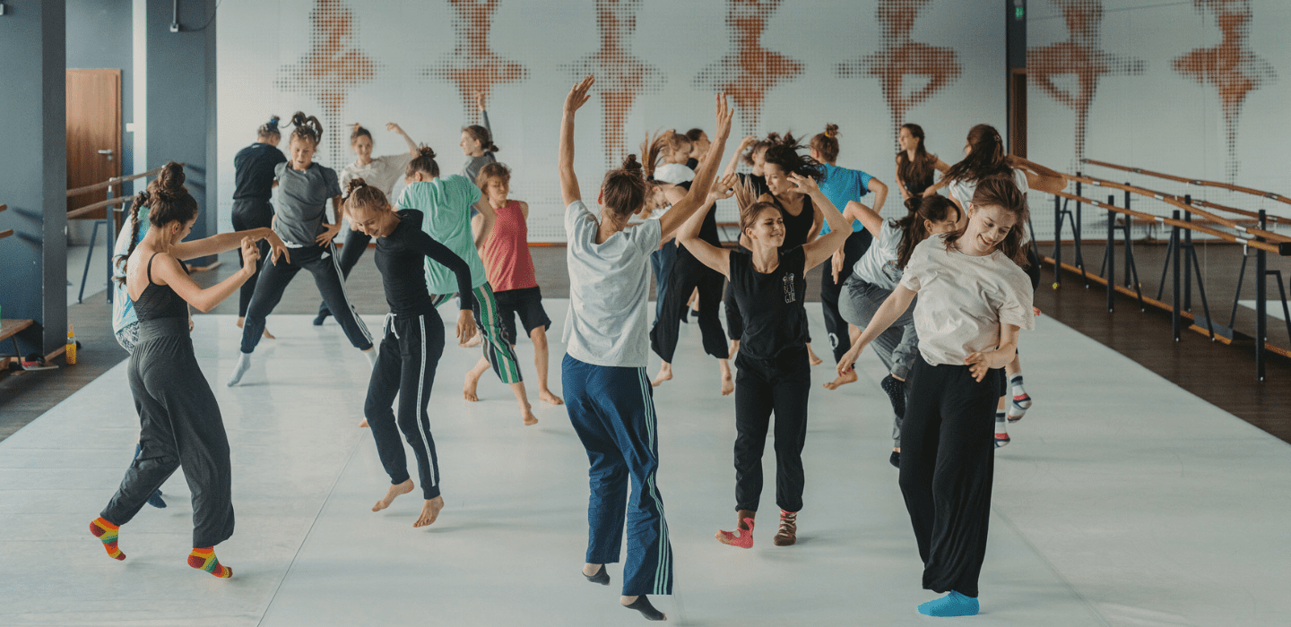 Grupa tańczących kobiet i mężczyzn w kolorowych ubraniach. Trening odbywa się w sali baletowej.
